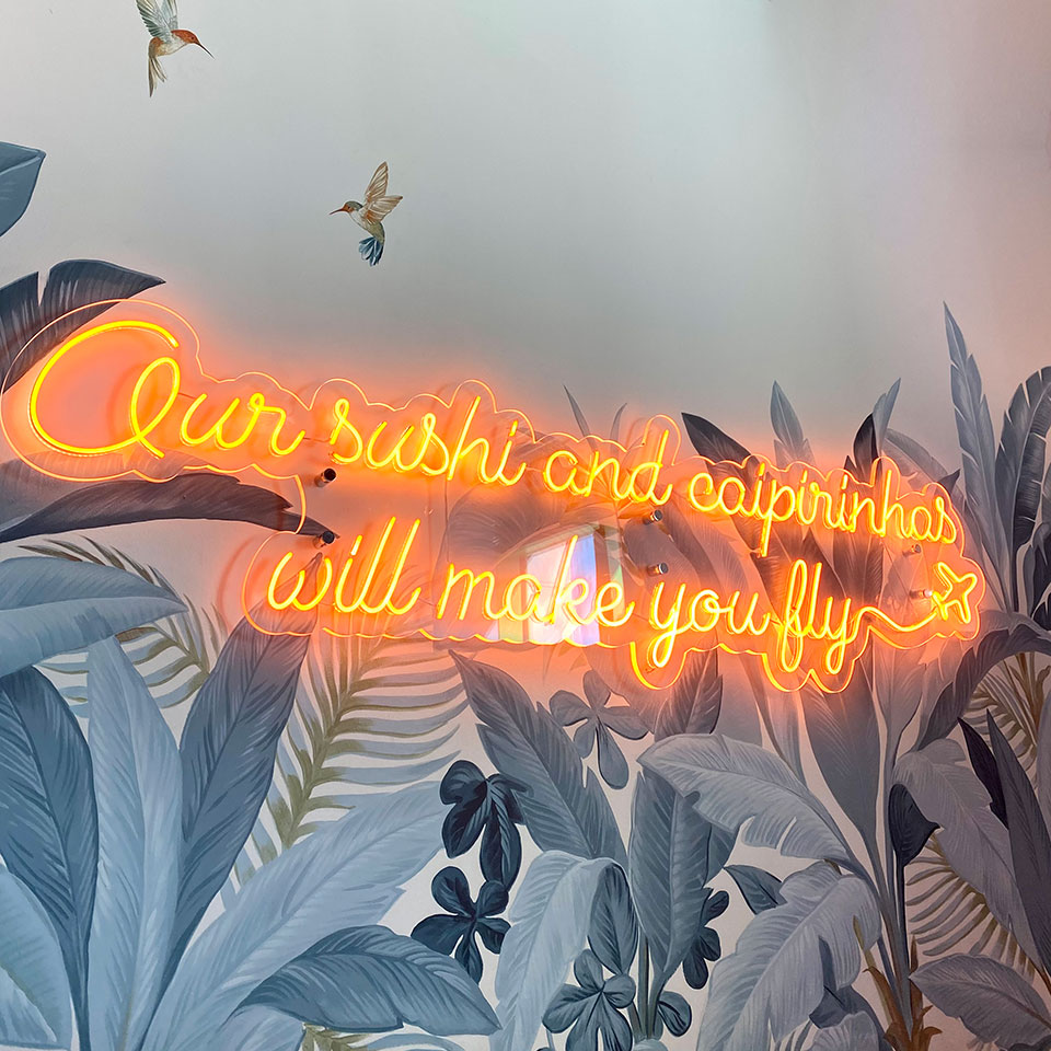 Parete del locale con insegna luminosa &ldquo;Our sushi and caipirinhas will make you fly&rdquo;.